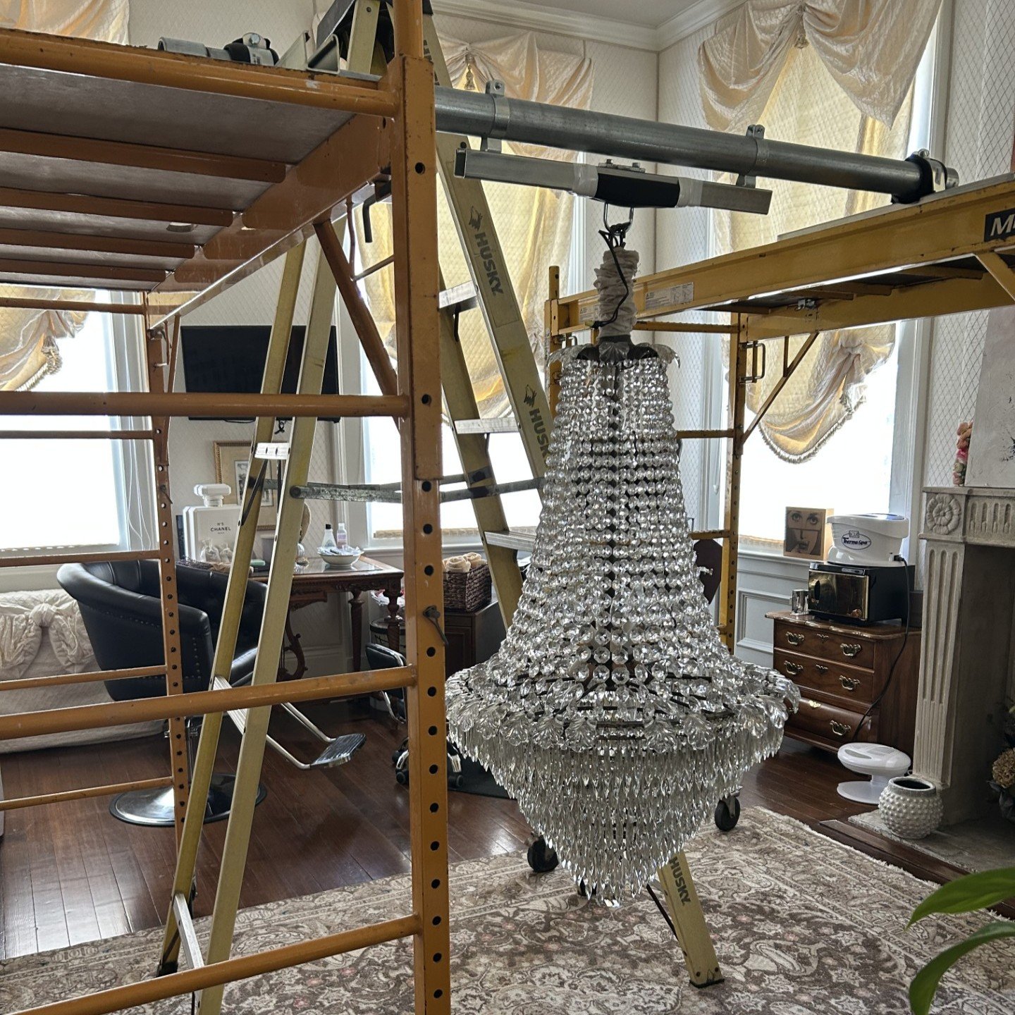 Large crystal chandelier suspended beneath lifting equipment beside scaffolding during installation.